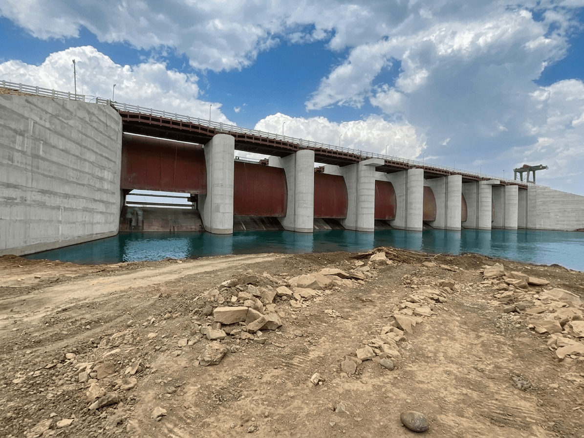Ground-level view along dam face with large red radial gates between white concrete piers and turquoise water